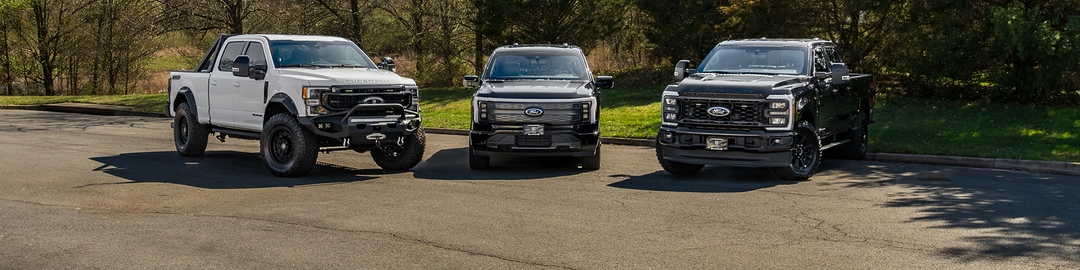  Three Alpine Armoring vehicles on a lot: a gray pickup truck with off-road mods, an emergency response vehicle with red and blue lights, and a black armored SUV with military features.