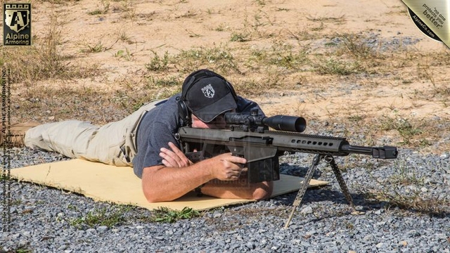 A person lies prone on a yellow mat, aiming a long-barrel rifle with a scope at an outdoor shooting range. The ground is a mix of small rocks and patches of dry grass.