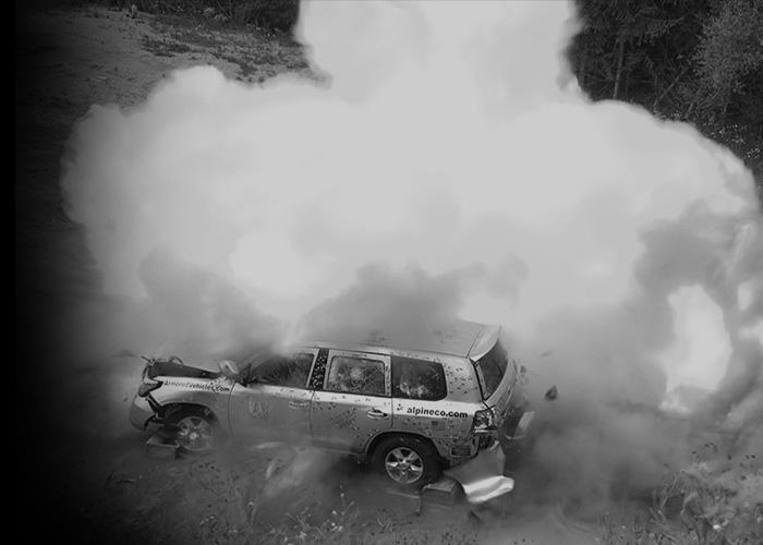 A white SUV, marked with "alpineco.com," is heavily damaged and surrounded by a large cloud of smoke, suggesting an explosion or collision in a wooded area.