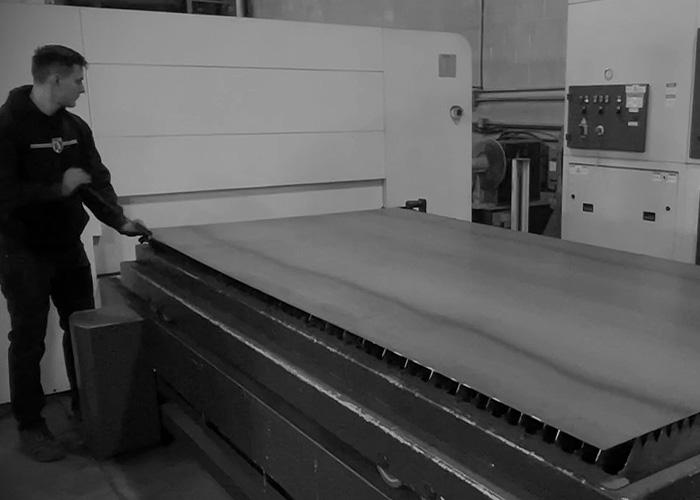 An Alpine Armoring employee operates a large industrial machine handling a flat metal sheet, inside a factory setting.