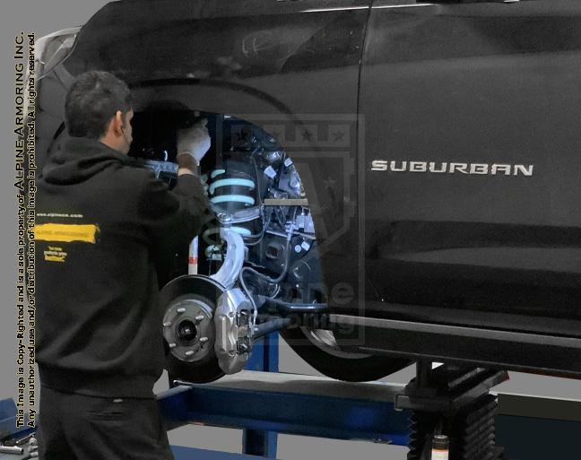 A technician at Alpine Armoring working on the suspension system of a black Chevrolet Suburban. The vehicle is lifted on a hydraulic platform, showcasing the detailed work involved in armoring and upgrading the vehicle's components.