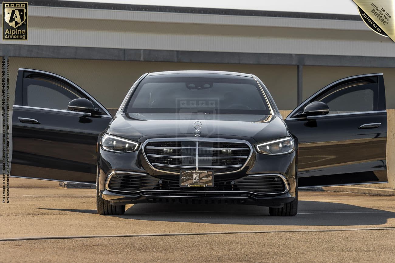 An armroed black Mercedes-Benz S580 sedan is parked with both front doors open, facing the camera. An Alpine Armoring logo is visible on a banner in the top left corner.