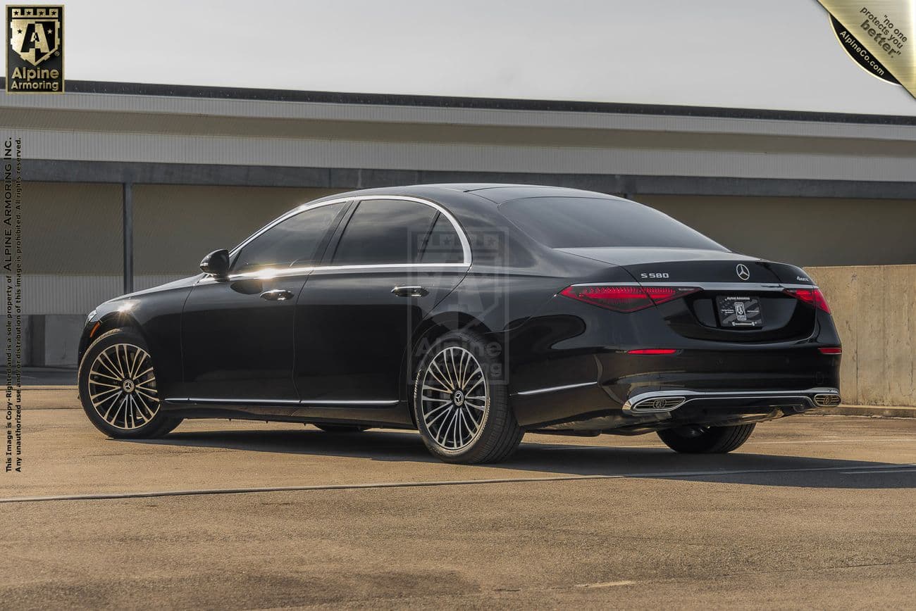 An armored black Mercedes-Benz S580 Executive sedan  is parked in a parking lot, featuring sleek lines and a polished finish. The background shows a building with a flat roof.