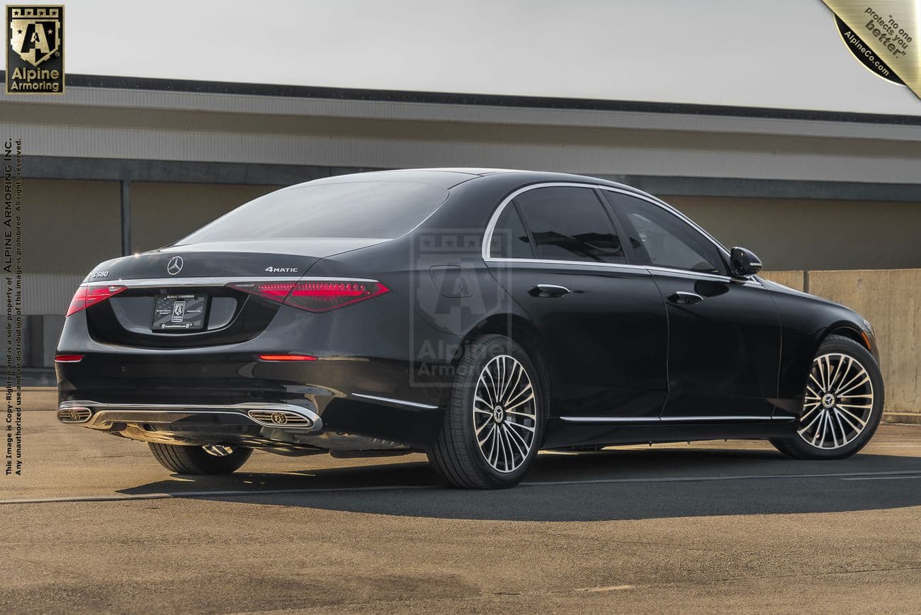 A black, armored Mercedes-Benz S580 sedan is parked on a concrete surface next to a building. A "Alpine Armoring" logo and details are imprinted on the image.