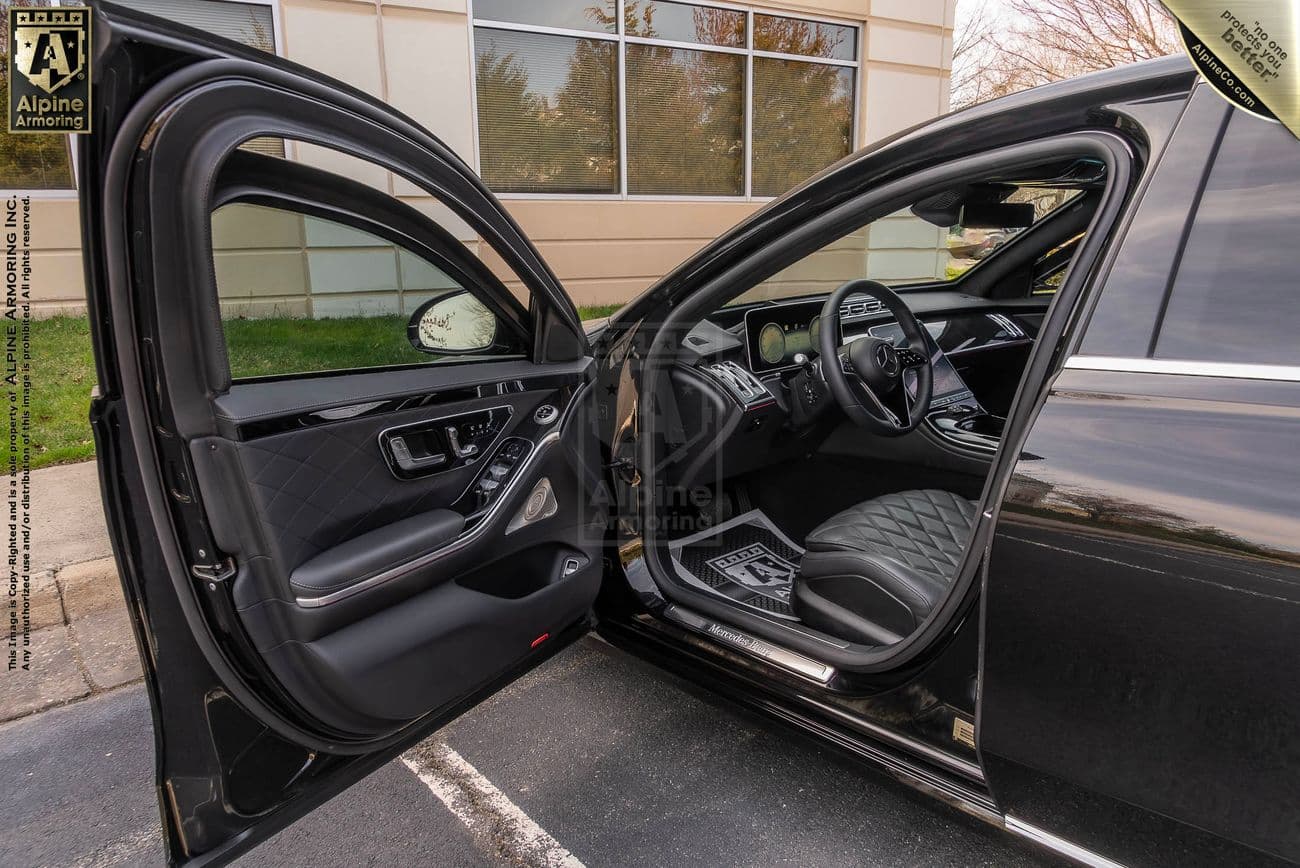 Open front driver-side door of a black armored Mercedes-Benz S580 sedan showing the interior with quilted leather seats and advanced dashboard technology. The car is parked in a lot beside a building with large windows.