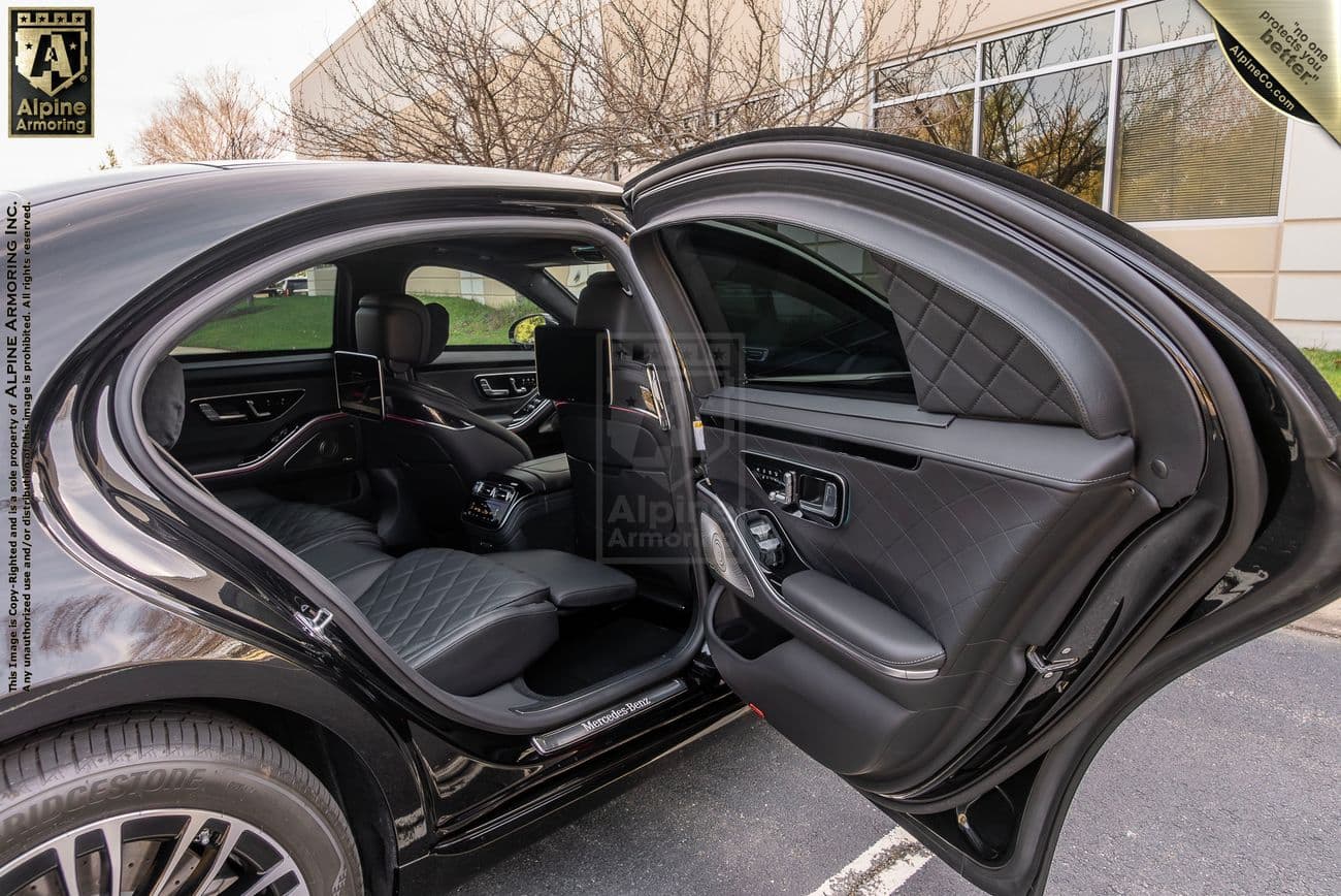 The open back door of an armored Mercedes-Benz S580 luxury car, revealing a spacious interior with leather seats and a screen attached to the back of the front seat.