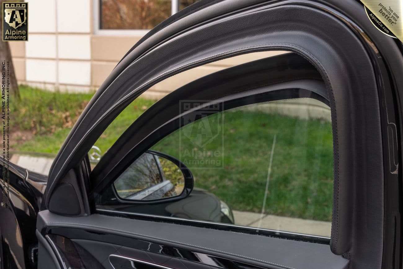 View of a car's open door window with bulletproof glass showcasing Alpine Armoring branding. The background includes a green lawn, windows, and the side mirror reflecting some of the surroundings.