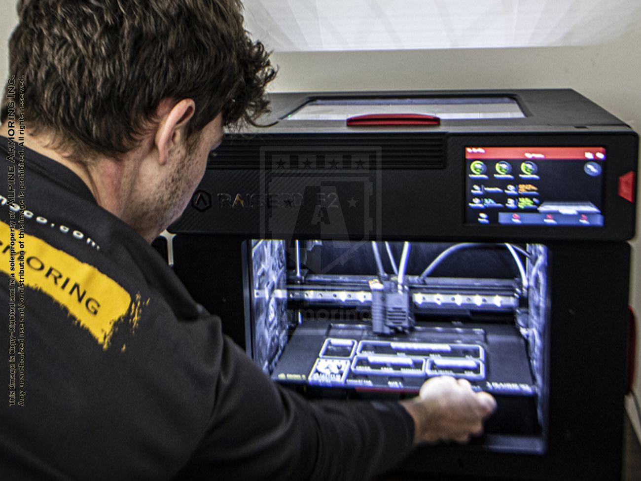 An Alpine Armoring employee operates a Raise3D E2 3D printer, adjusting the machine's interior while a 3D model is being printed.