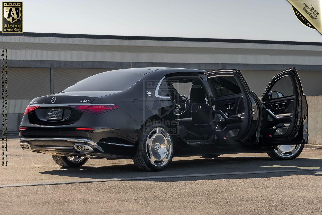 A black armored Mercedes-Benz S 580 Maybach sedan is parked in a lot with both rear doors open, showing the interior. The car is detailed by Alpine Armoring.