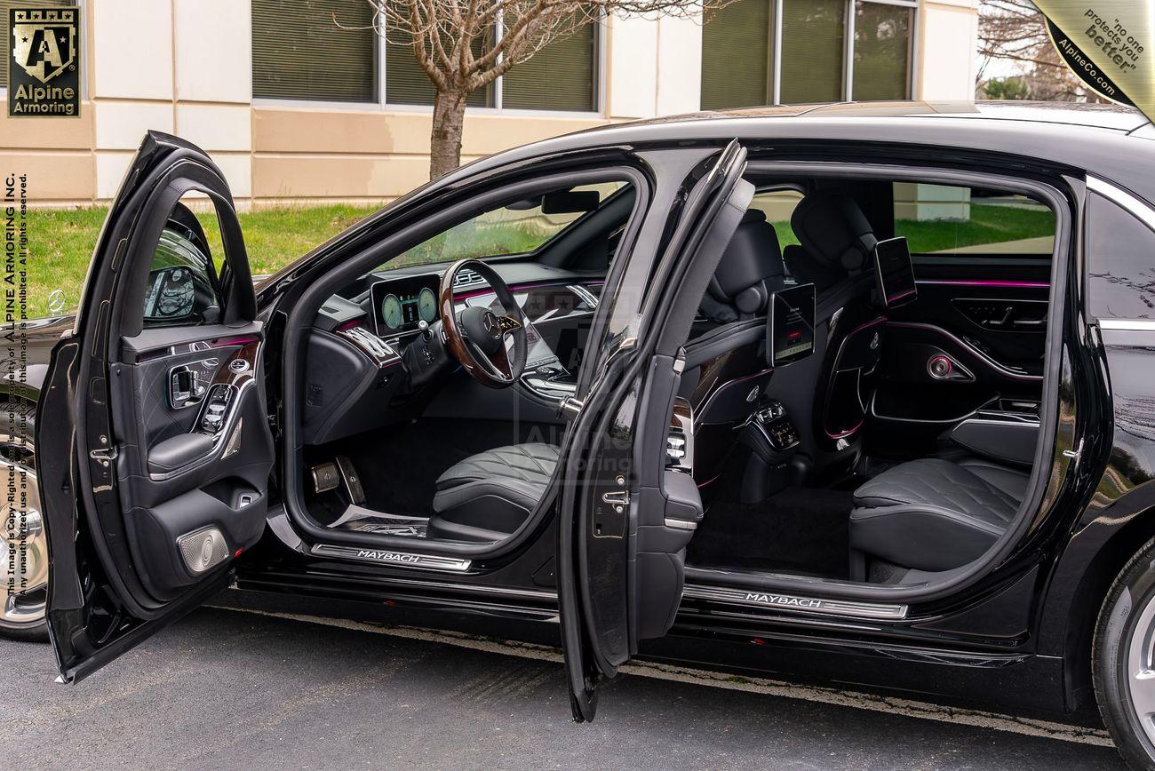 A black armored Mercedes-Benz S 580 Maybach sedan with all its doors open, revealing an interior with leather seats, digital dashboard, and ambient lighting. The car is parked in front of a building.