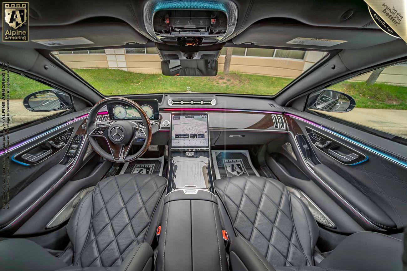 Interior view of an armored Mercedes-Benz S 580 Maybach sedan showcasing a sleek dashboard, central touchscreen display, and leather quilted seats. Steering wheel features a prominent car logo, with ambient lighting lining the doors.