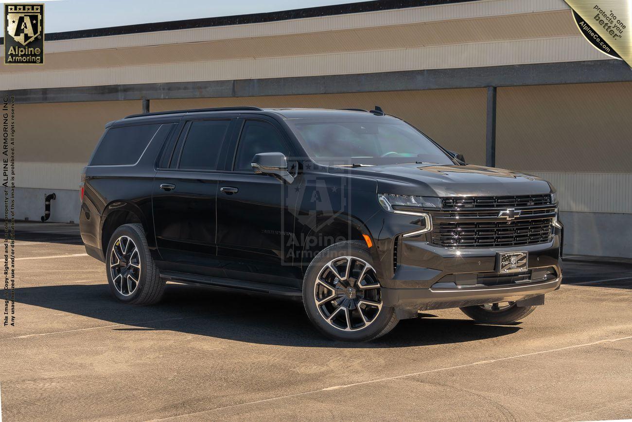 A black armored Chevrolet Suburban LT SUV with dark tinted windows is parked outdoors in front of a building. The SUV has black alloy wheels and is photographed from the front/side angle.