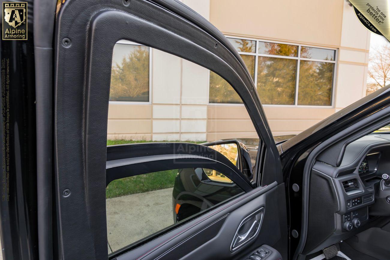 A partially open driver's side door of a black Chevrolet Suburban LT vehicle reveals the interior, showcasing the car's bulletproof glass window, mirror, and part of the dashboard. The car is parked next to a beige building with windows.