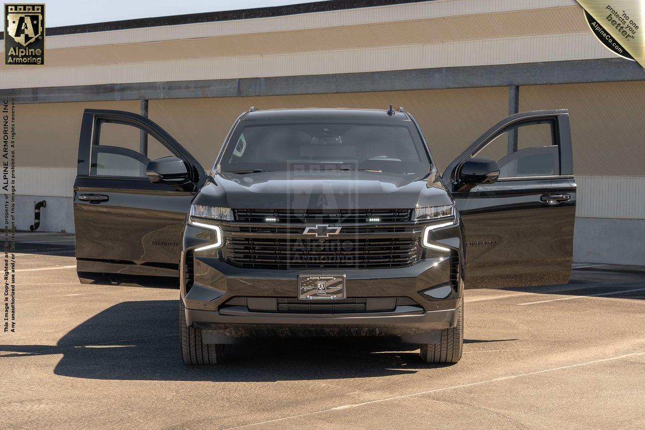 Front view of a black armored Chevrolet Suburban LT SUV with both front doors open, parked in a lot with a building in the background.