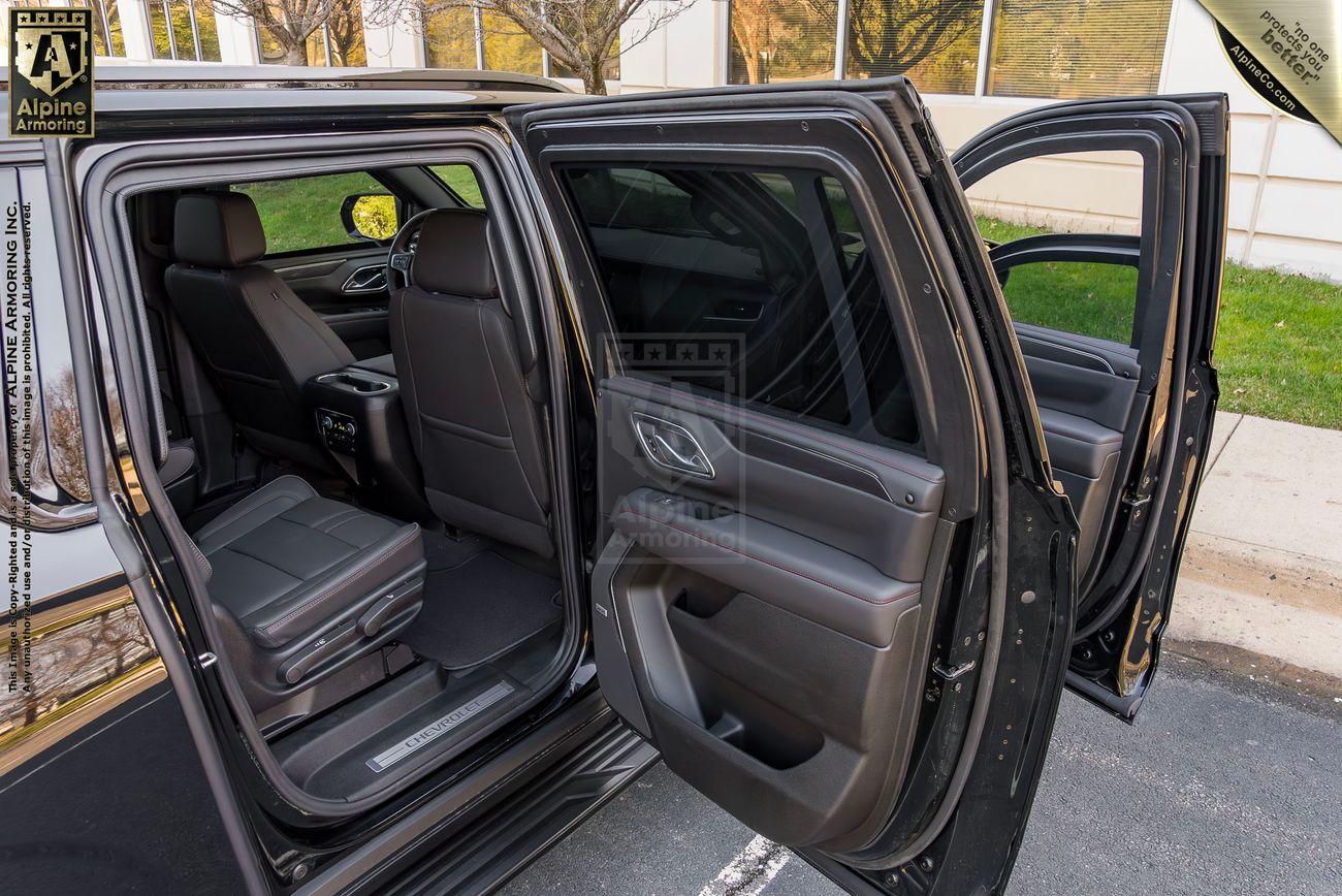 The interior view of an open black armored Chevrolet Suburban LT SUV door showing black leather seats, a center console, and a clean, spacious rear seating area. The window of fhe front door is rolled partially down, revealing bulletproof glass.