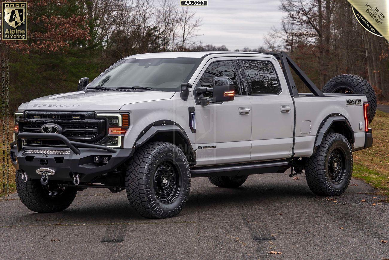 A white heavy-duty Mastiff truck with off-road modifications, including all-terrain tires, a front bumper guard, and a rear roll bar with a mounted spare tire, parked on a road with trees in the background.