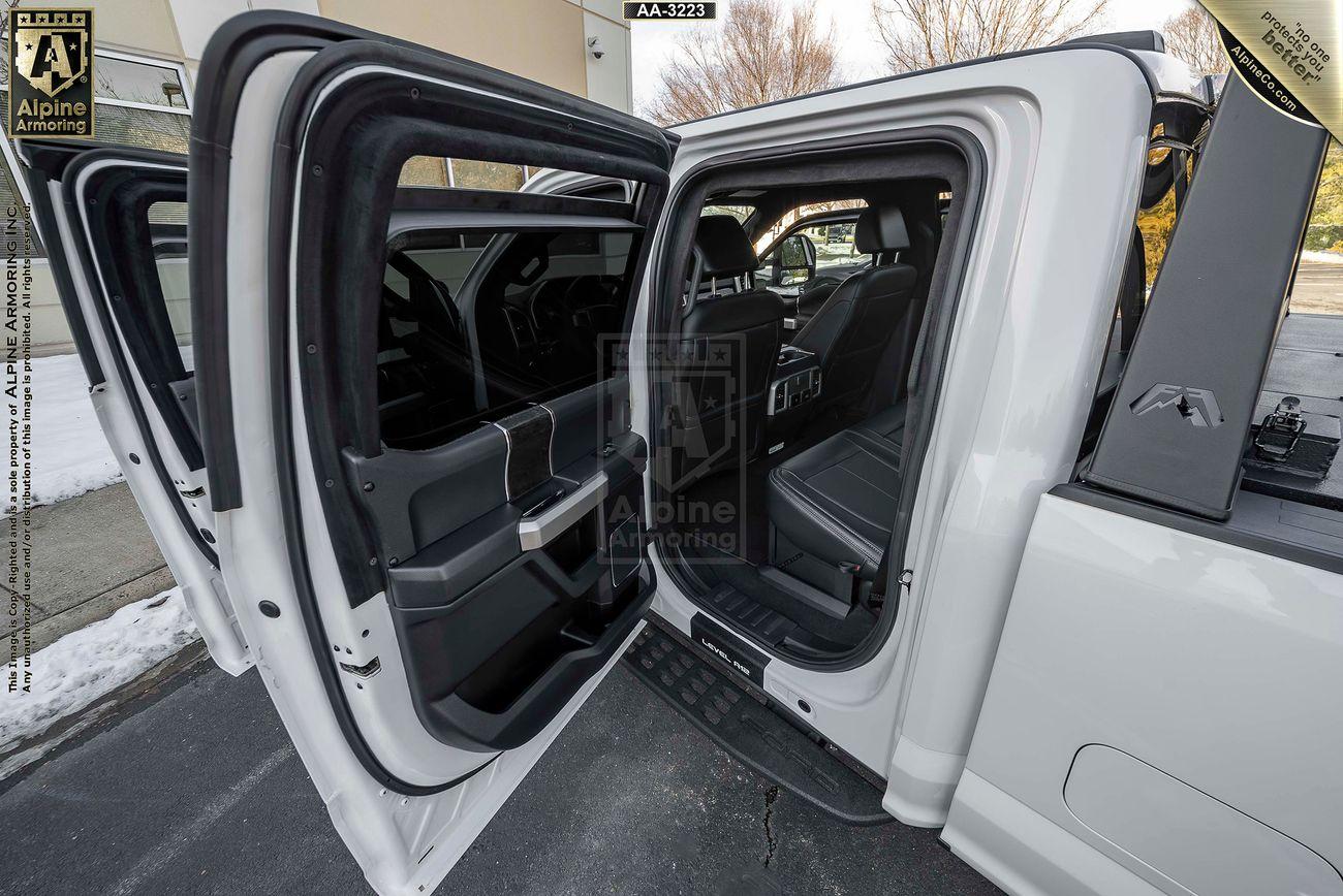 Open rear doors of a white Mastiff revealing two rows of black leather seats and interior features. The outside environment appears to be a parking area on a clear day.