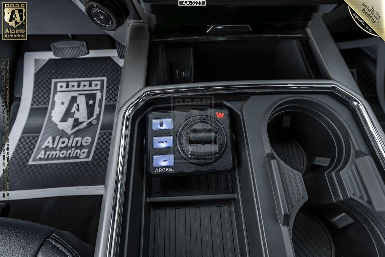 Close-up of a Mastiff's center console showing three labeled buttons, a dial, and two cup holders. A branded Alpine Armoring floormat is partially visible underneath.