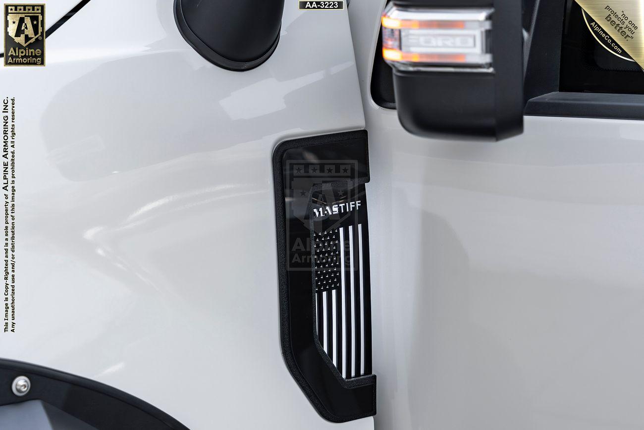 Close-up of a part of a white Mastiff, showing a black American flag decal with a blue stripe between the front fender and side mirror. Text "ALPINE ARMORING" is partially visible on the left.