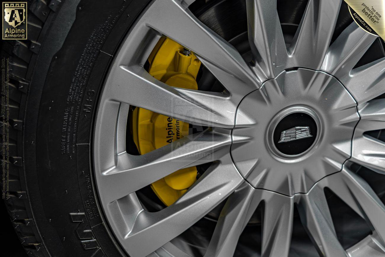 A close-up of a vehicle wheel, showcasing an Alpine Armoring yellow brake caliper through the alloy wheel spokes.