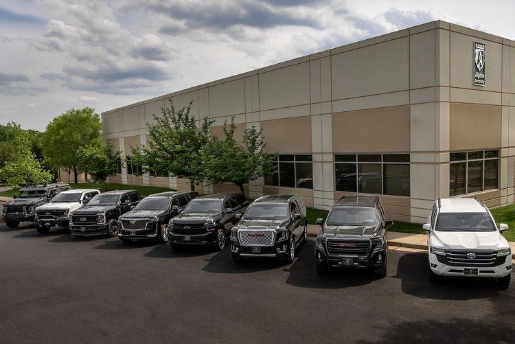 Eight Alpine Armoring SUVs and trucks, in colors ranging from black and gray to white, are parked in front of a beige building with a logo. Green trees surround the Alpine Armoring building under a cloudy sky.