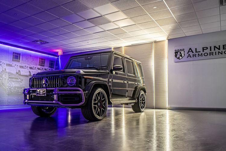 A black Mercedes-Benz SUV is parked in the Alpine Armoring showroom. Above it, signs read "No one protects you better" and "Alpine Armoring." The room has a tiled floor and a drop ceiling with fluorescent lights.