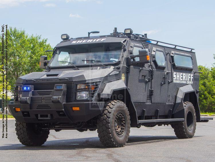 An armored sheriff's Pit-Bull vehicle labeled "SHERIFF" is parked on a paved surface with trees visible in the background. The vehicle is equipped with off-road tires and various lights.