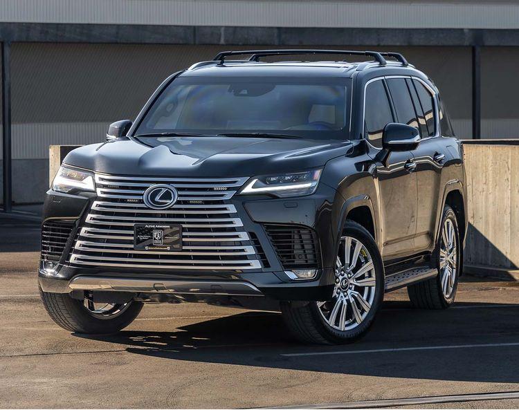 A dark-colored Lexus LX 600 Ultra Luxury SUV is parked in a parking lot near a concrete barrier, featuring a prominent front grille and roof rails.