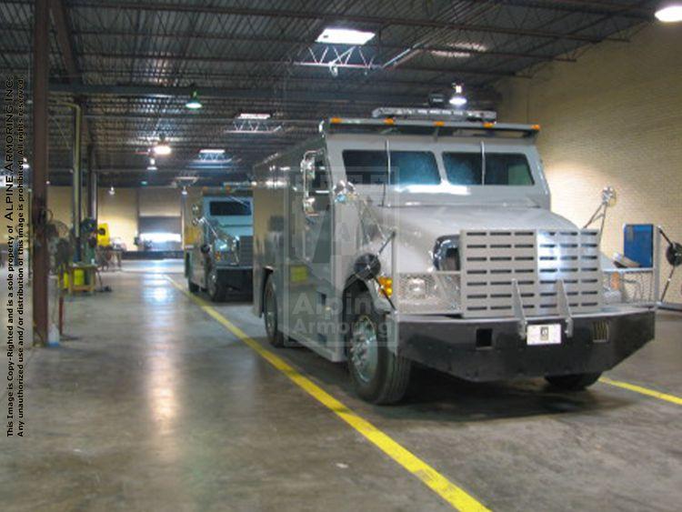 Two armored CIT trucks parked inside a well-lit warehouse with concrete floors and exposed ceiling beams. The vehicles are positioned in aligned parking spaces with yellow lane markings.