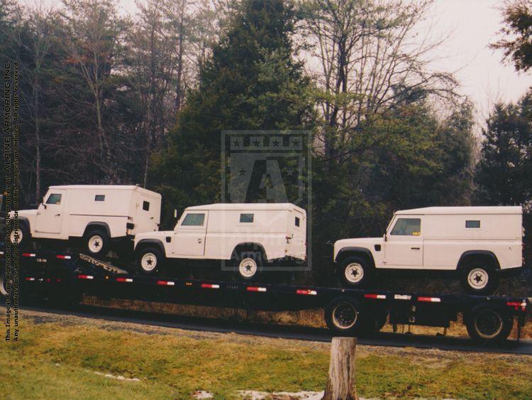 Three white SUVs are loaded onto a flatbed truck, parked on a road with a backdrop of trees.