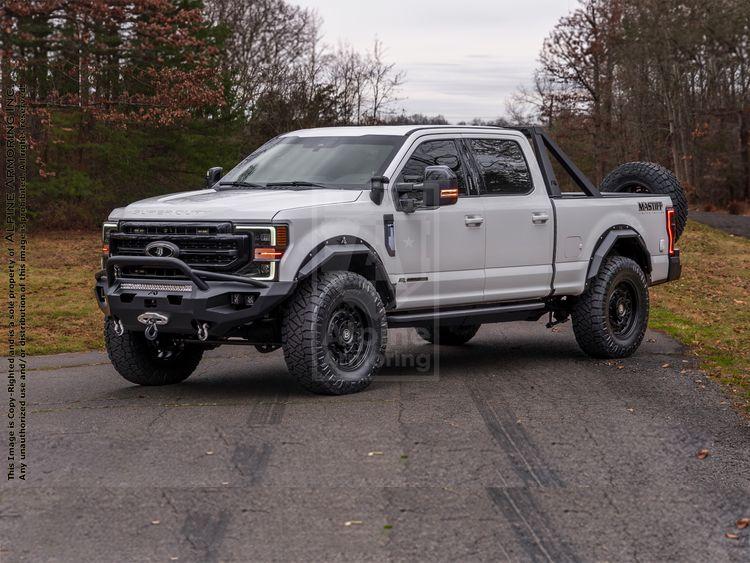 A white mastiff truck with rugged off-road tires is parked on a paved road in a wooded area, with trees and patches of grass visible in the background.