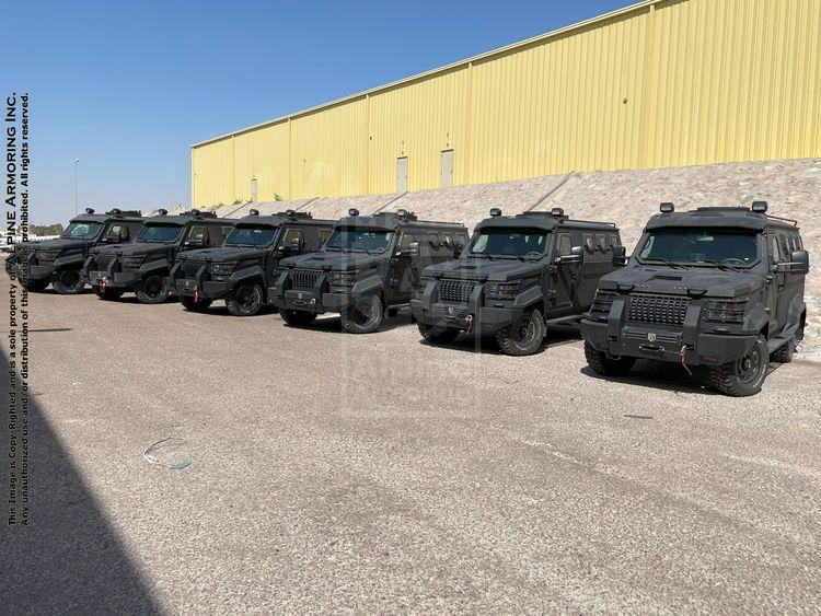 A row of black armored Cudas is parked outside a large yellow industrial building on a sunny day.