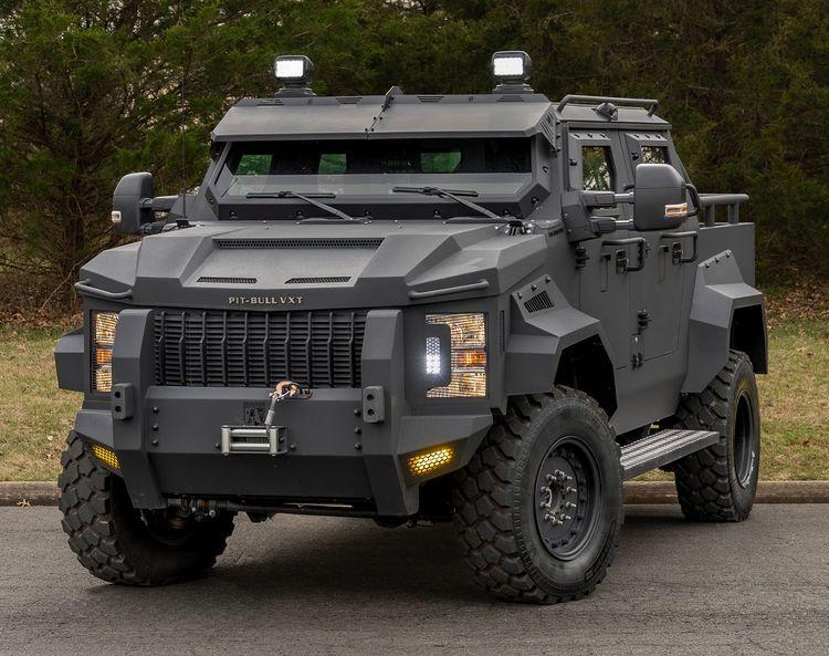 A black armored Pit-Bull VXT with rugged tires and external lights is parked on a paved surface with trees in the background.