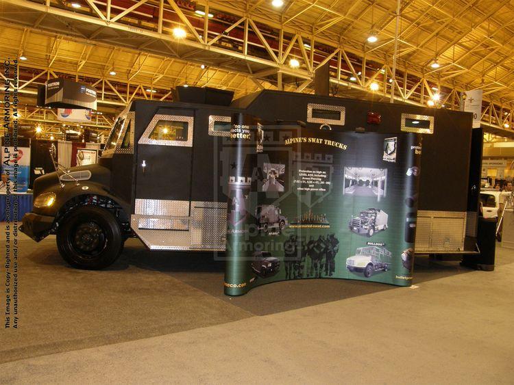 A black armored Alpine Armoring Bulldog displayed indoors at an exhibition, showcasing images and features of SWAT trucks on a poster board attached to its side.