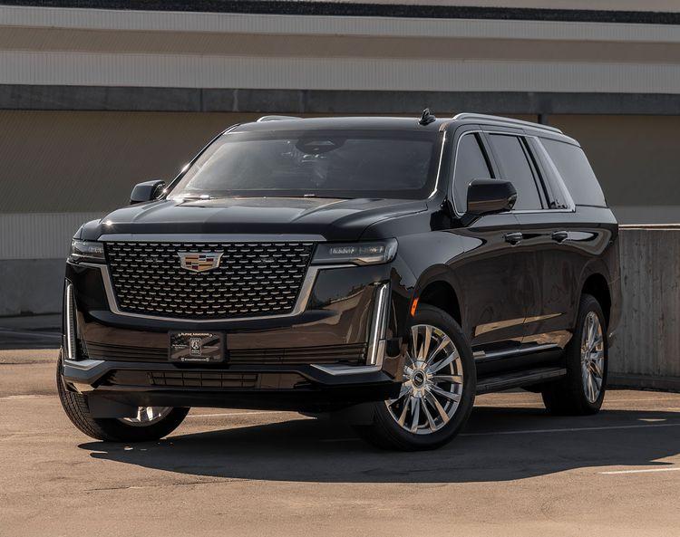 A black armored Cadillac Escalade ESV  with a Cadillac emblem on the grille is parked in an outdoor area on a sunny day.