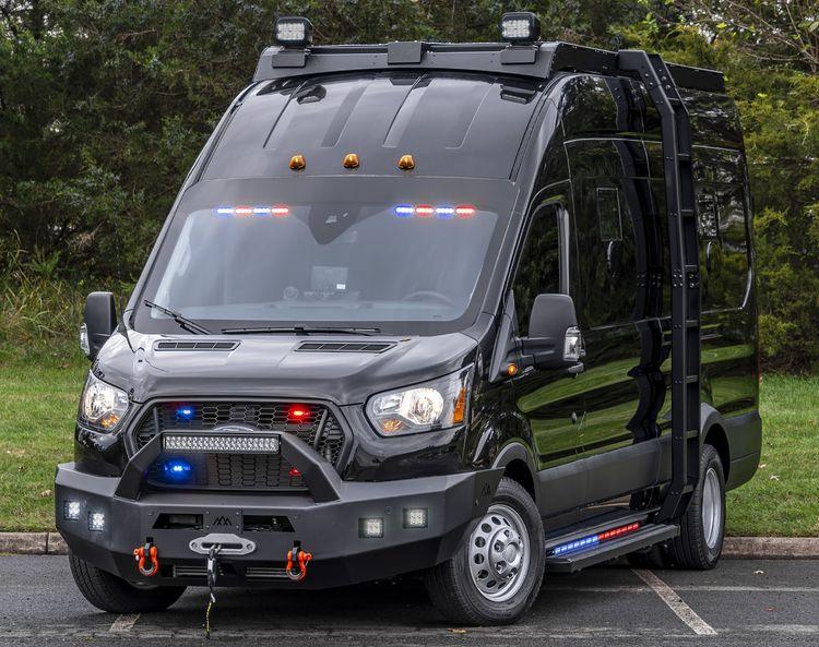 Black, armored Ford Transit Pointer Van with various emergency lights and sirens, parked in front of a grassy area with trees in the background.