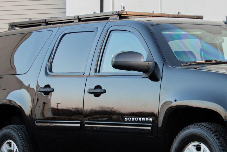 A black SUV with tinted windows, marked “Suburban” on the side, is parked in front of a beige building. The vehicle has a roof rack installed. The image captures the front and side of the SUV.