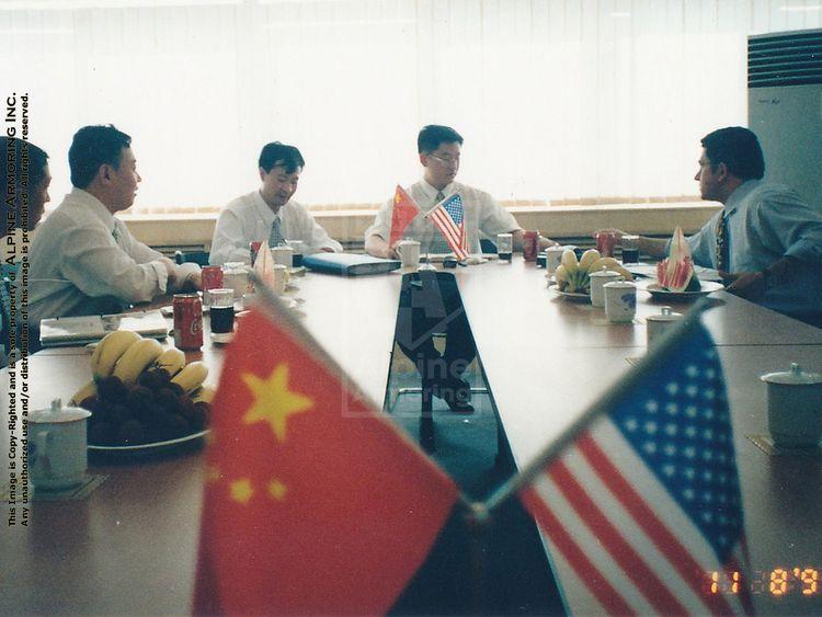 Four men are seated at a conference table, engaged in a meeting. Chinese and American flags are displayed on the table, along with refreshments including bananas and soda cans.