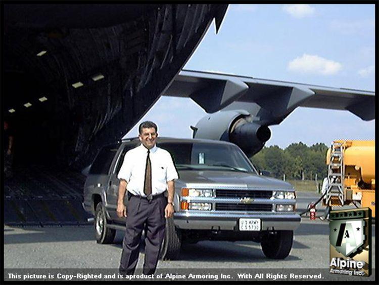 A man stands in front of a gray SUV parked near the open rear cargo bay of a large aircraft. He is dressed in a white shirt and tie.