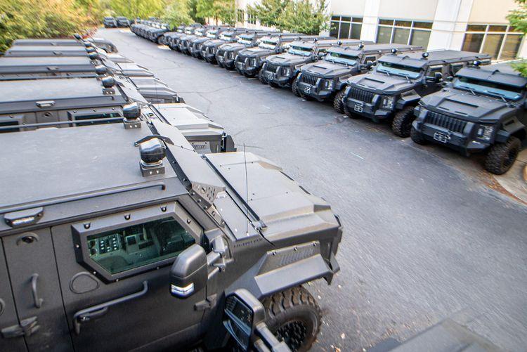 A large fleet of black Alpein Armoring SWAT vehicles are parked in rows outside a building with white and beige walls. The vehicles are aligned diagonally, occupying most of the visible parking lot. Trees and greenery can be seen in the background.