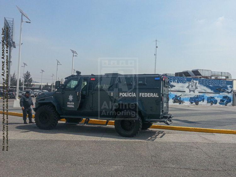 A black armored police vehicle marked "Policía Federal" is parked on a street near a mural depicting police operations. A few individuals in uniform stand nearby.