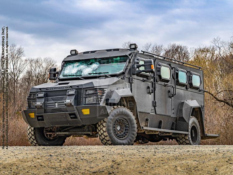 An armored Pit-Bull vehicle with a bullet-damaged windshield is parked on a dirt road. Trees and bushes are in the background.