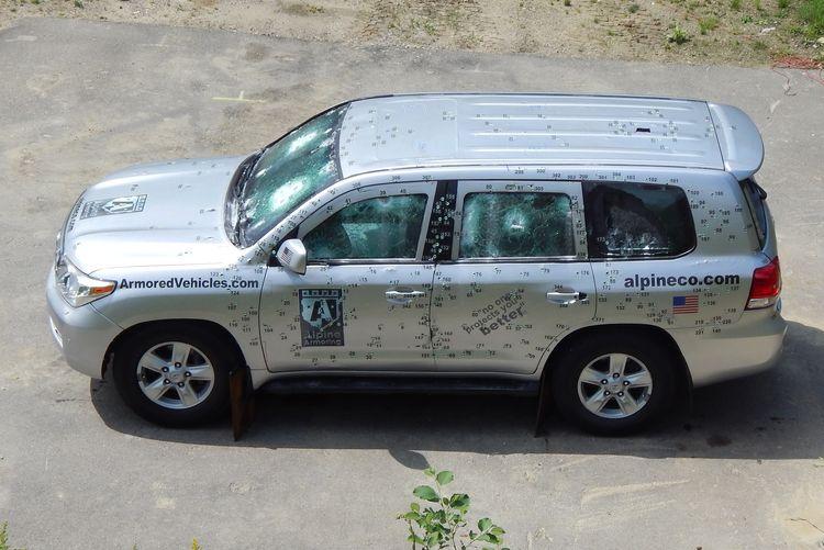 A silver armored SUV riddled with bullet holes is parked on a concrete surface. The vehicle has several markings, including "ArmoredVehicles.com" and "alpineco.com" on its sides, and has undergone a ballistic test.