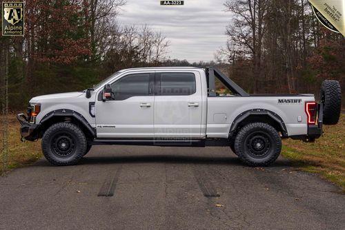 Driver side view of a white Mastiff truck with black trims and rugged tires is parked on a road surrounded by trees. It features a mounted spare tire on the back and a roll bar over the bed.