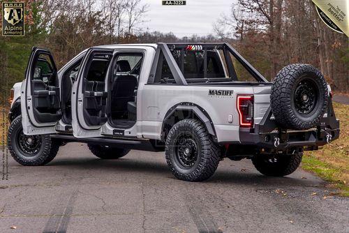 A modified Mastiff with open driver side doors, large tires, and a mounted spare tire on the back on a paved road with trees in the background.