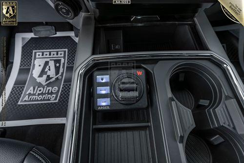 Close-up of a Mastiff's center console showing three labeled buttons, a dial, and two cup holders. A branded Alpine Armoring floormat is partially visible underneath.