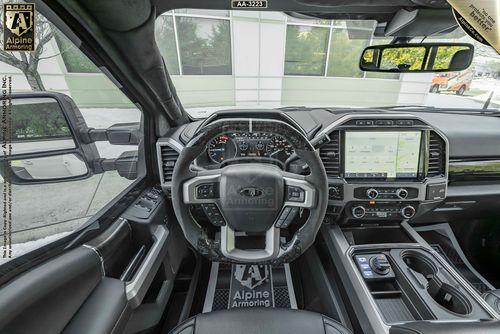 Interior view of a Mastiff's driver cockpit featuring a steering wheel with the Alpine Armoring logo, a large touchscreen display, and various control buttons and dials on the dashboard and center console.