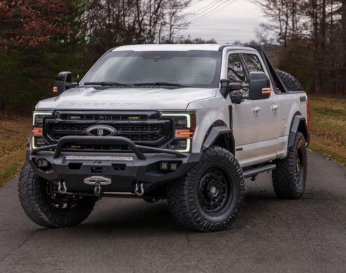 A silver lifted Ford F-350 Mastiff with off-road tires is parked on a paved road surrounded by trees on an overcast day.