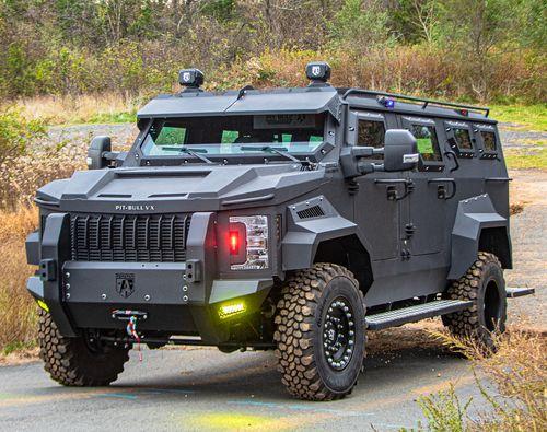 An armored Pit-Bull VX with a rugged design and off-road tires is parked on a paved road, surrounded by vegetation.